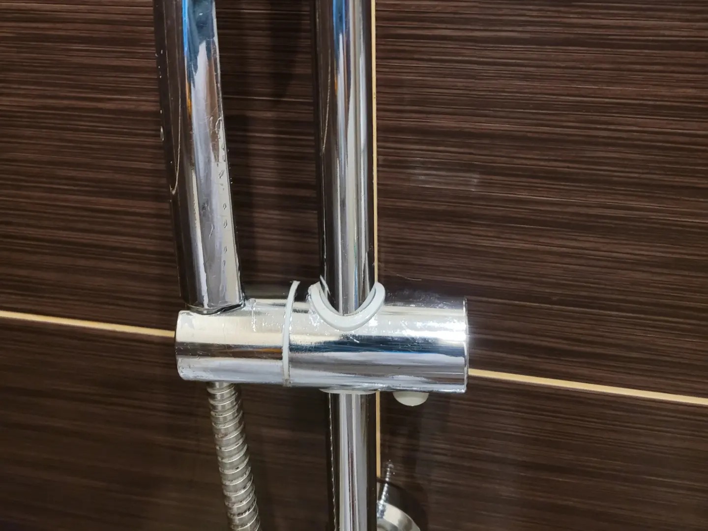 Błyszcząca, chromowana bateria prysznicowa z widocznymi kroplami wody na tle ciemnobrązowej ściany z poziomymi paskami. Detal po czyszczeniu.