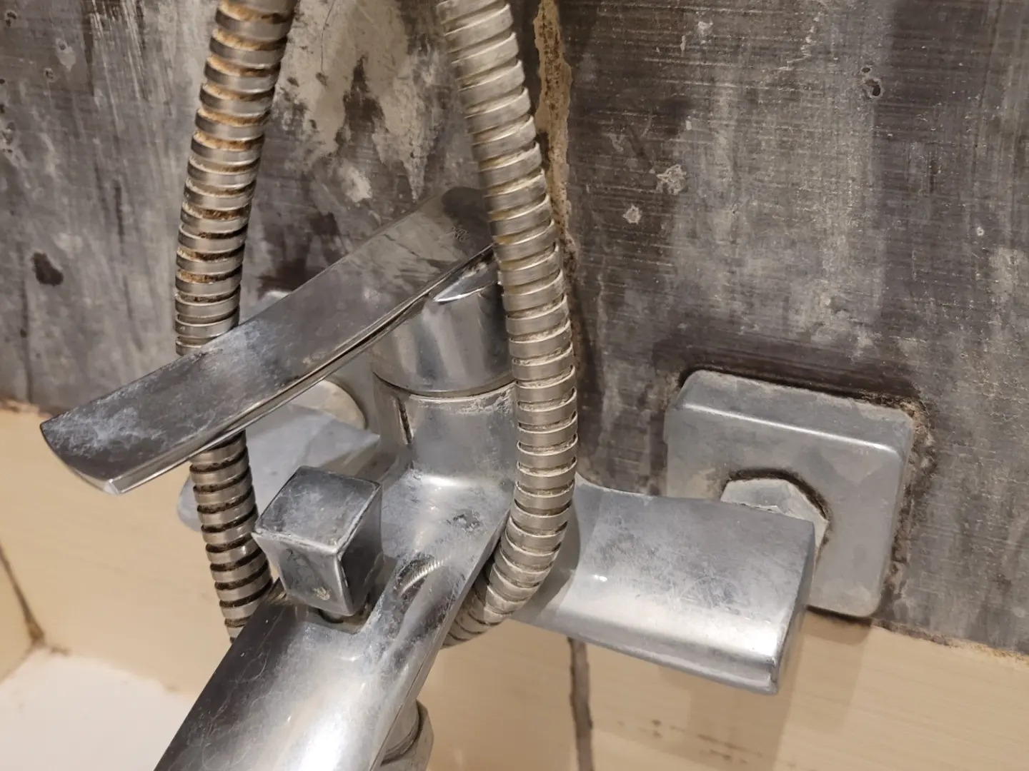 Z bliska: zanieczyszczona, chromowana bateria prysznicowa z elastycznym wężem na tle szarej, zabrudzonej ściany. Widoczne osady i zacieki.