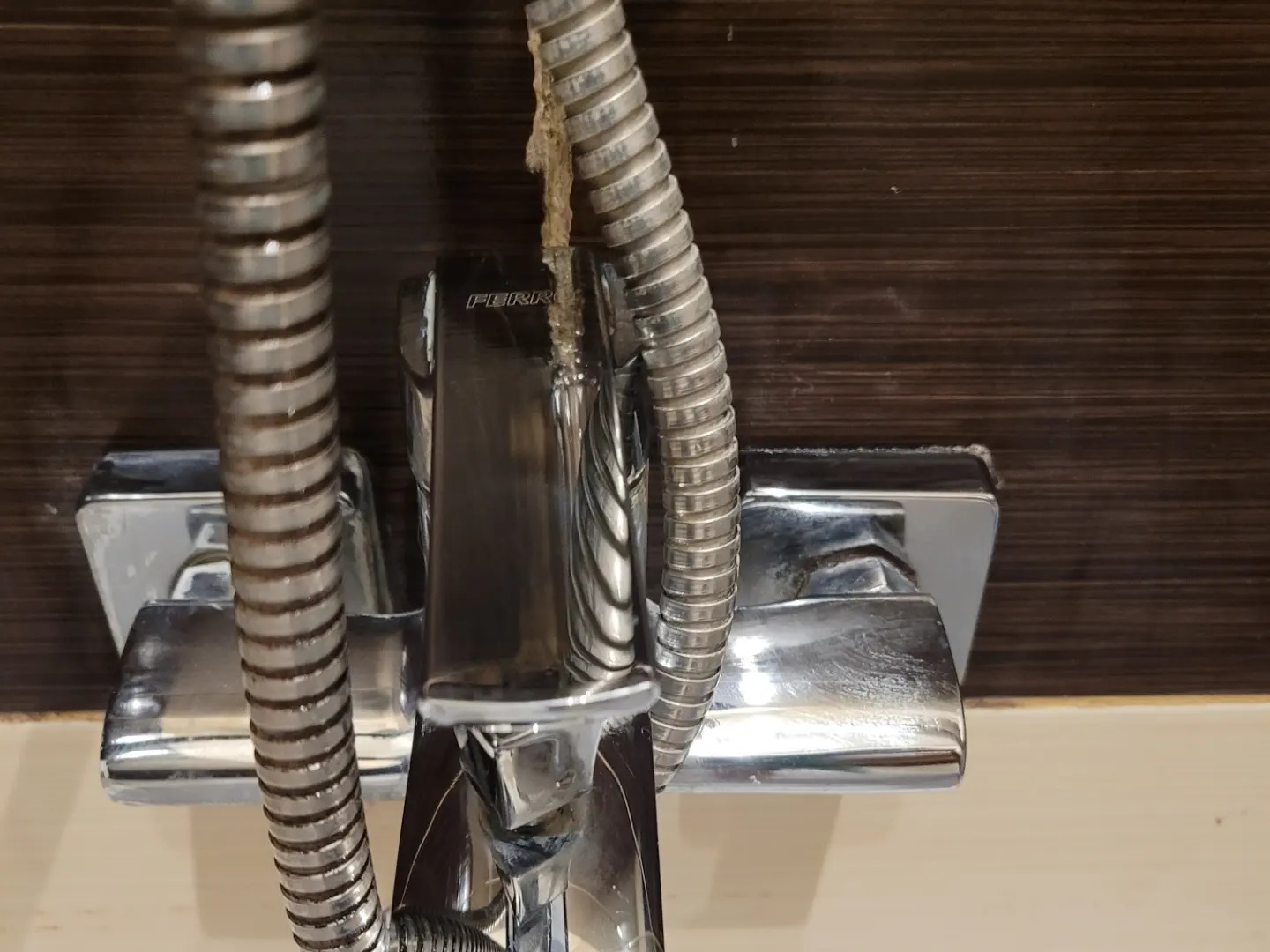 Zanieczyszczona, chromowana bateria prysznicowa z osadem i kamieniem, widoczna na tle ciemnej, drewnopodobnej ściany. Wąż prysznicowy z widocznymi zabrudzeniami.