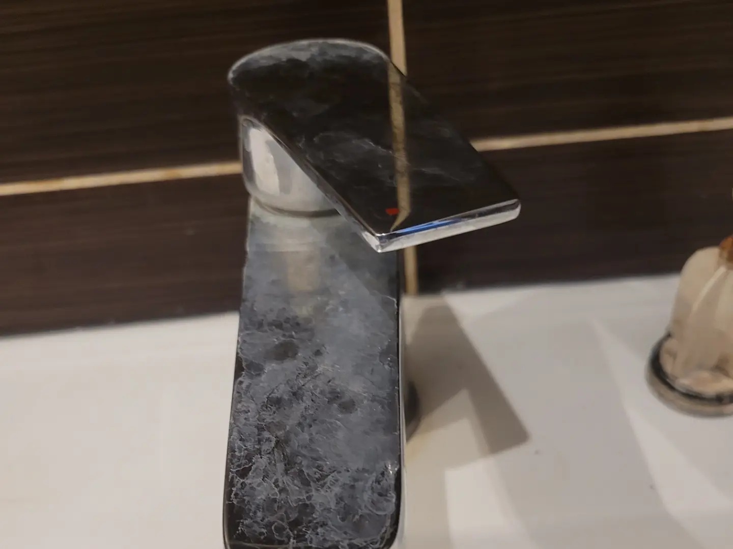 Z bliska: chromowana bateria umywalkowa z wyraźnym osadem kamiennym na powierzchni, na tle brązowych płytek i białej umywalki. Widoczne ślady użytkowania.