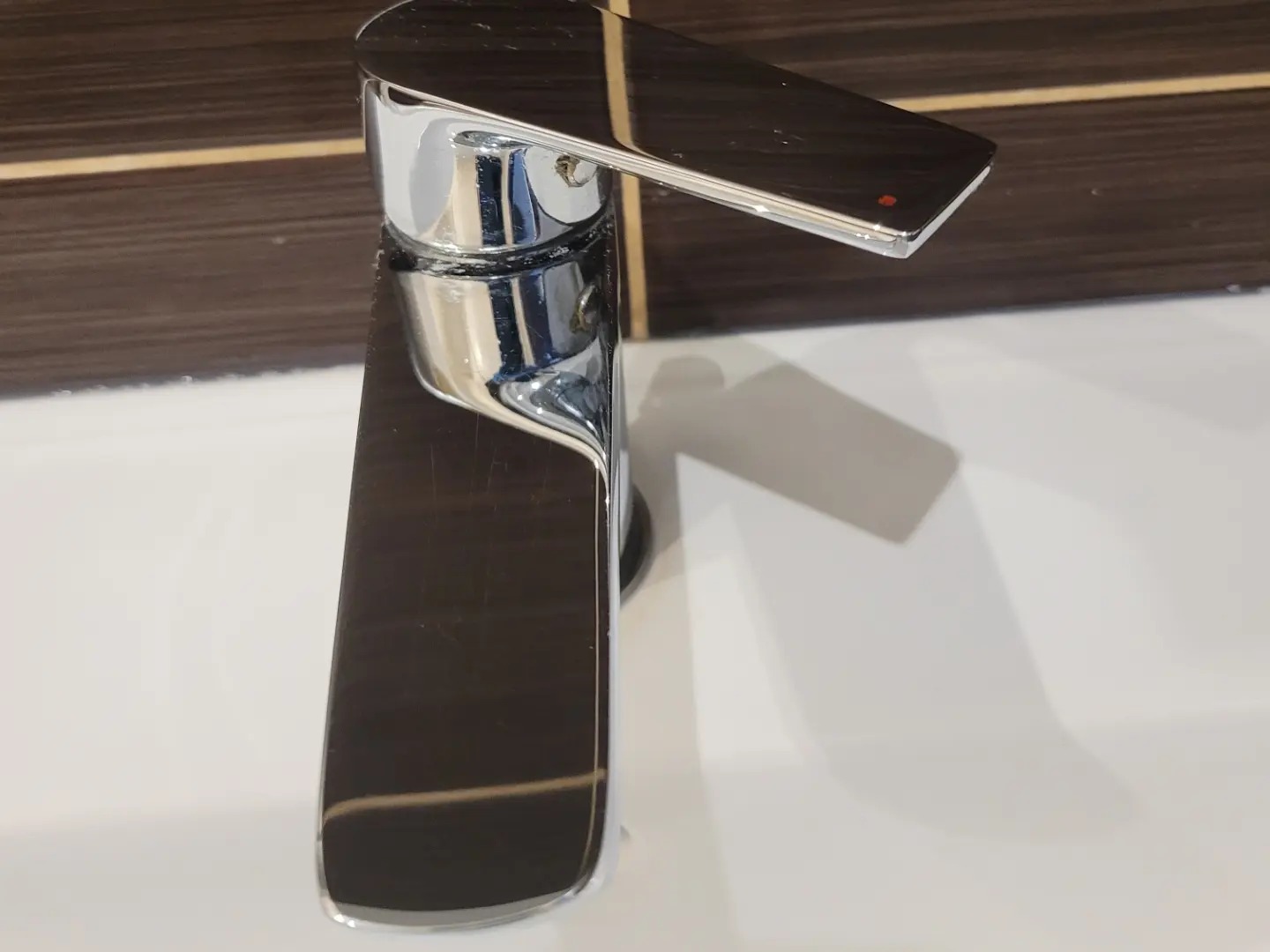 Błyszcząca, chromowana bateria umywalkowa z czerwoną kropką na dźwigni, osadzona na białej umywalce z ciemnobrązowym panelem w tle. Widoczne ślady użytkowania.