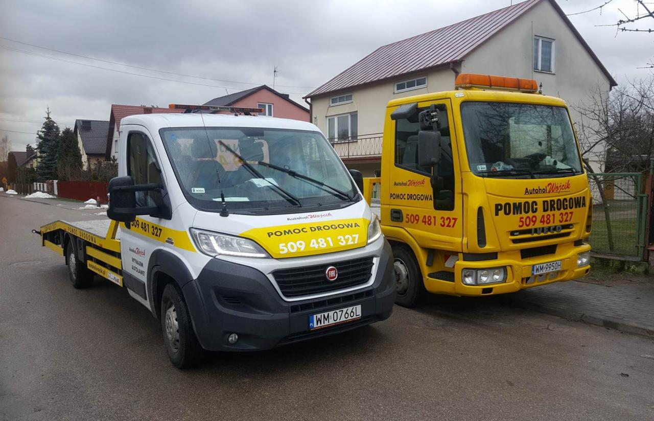 Dwie ciężarówki pomocy drogowej zaparkowane na ulicy przed budynkami mieszkalnymi, jedna biała Fiat Ducato z żółtymi elementami, druga żółta Iveco, obie z napisem 'Pomoc Drogowa' i numerem telefonu.