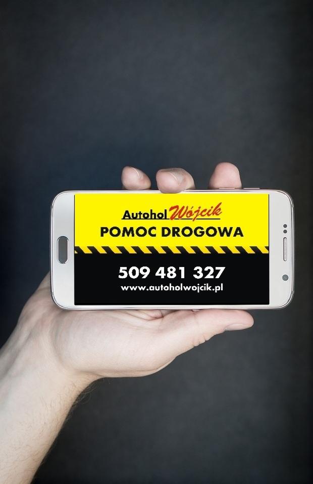 Smartfon w dłoni prezentujący reklamę firmy Autohol Wójcik oferującej pomoc drogową z numerem telefonu i adresem strony internetowej na żółto-czarnym tle.