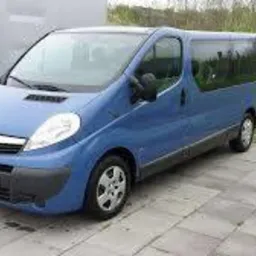 Niebieski Opel Vivaro, widok z przodu, zaparkowany na kostce brukowej, z widocznym fragmentem innego pojazdu z prawej strony.