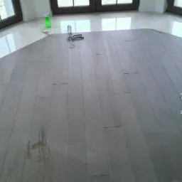 Jasnoszare drewniane deski podłogowe w trakcie montażu, widoczne narzędzia do klejenia i resztki materiałów, tło z marmurową posadzką i oknami.