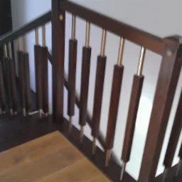 Klatka schodowa z ciemnobrązowymi drewnianymi balustradami z metalowymi, złotymi prętami, widok z góry na kontrastujące podłogi z jasnego i ciemnego drewna.