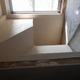 Wnętrze w trakcie prac wykończeniowych: ściany pokryte świeżą warstwą gładzi, widoczne ślady po szpachlowaniu, surowa podłoga i okno dachowe w górnej części kadru.