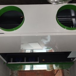 Good Live Solutions Szymon Nikiel - Zamontowany, biały rekuperator z zielonymi otworami wentylacyjnymi, widoczny z dołu, w surowym wnętrzu. Element instalacji wentylacyjnej w trakcie montażu.