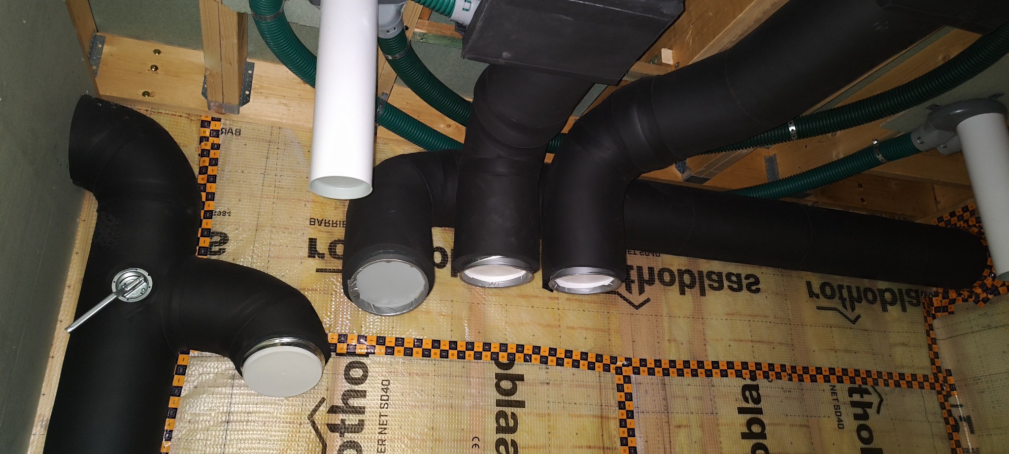 Czarno-białe rury wentylacyjne pod sufitem, z zielonymi przewodami karbowanymi. Widoczne mocowania i izolacja. Instalacja rekuperacji w trakcie montażu.