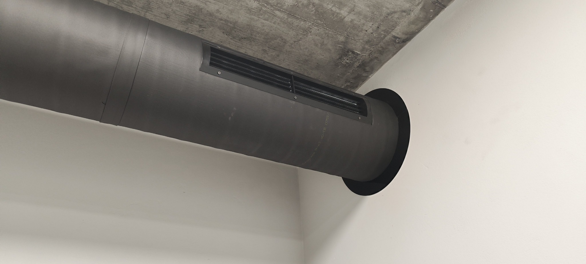 Szara rura wentylacyjna z kratką, zamontowana pod betonowym sufitem w rogu pomieszczenia z białymi ścianami. Widok z dołu.