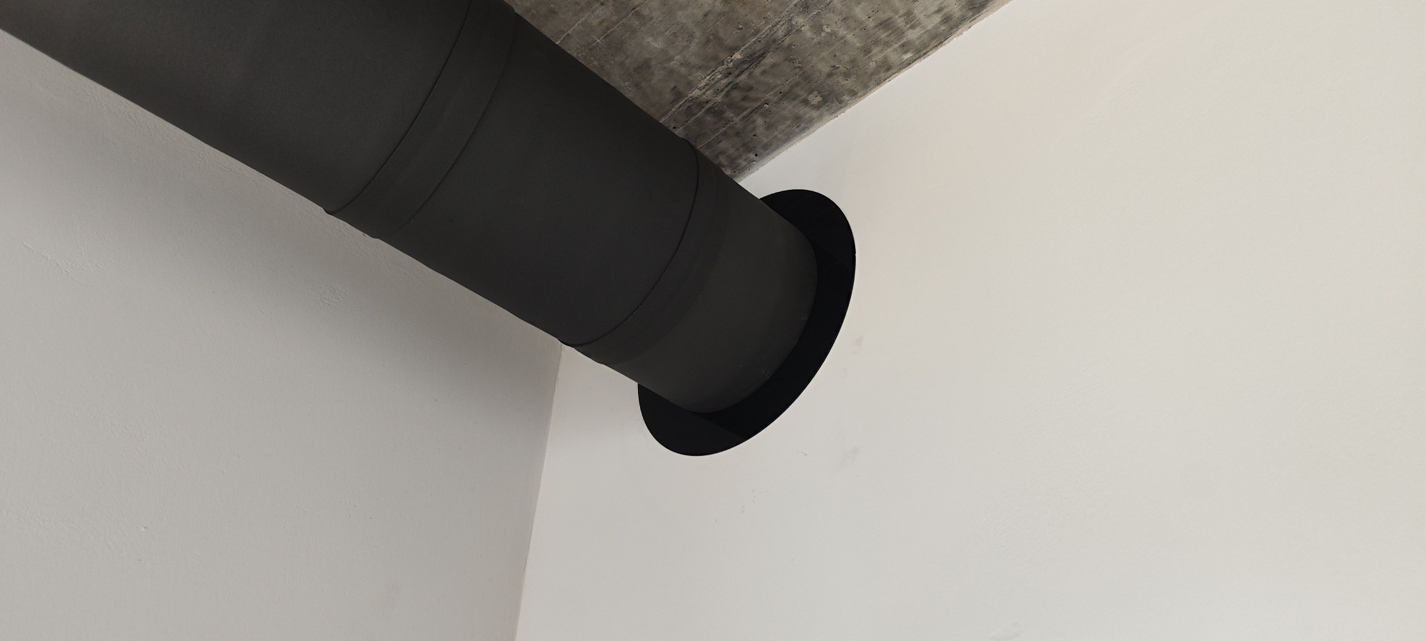 Czarny przewód wentylacyjny zamontowany pod betonowym stropem, w przejściu przez białą ścianę. Widoczne łączenie segmentów rury.