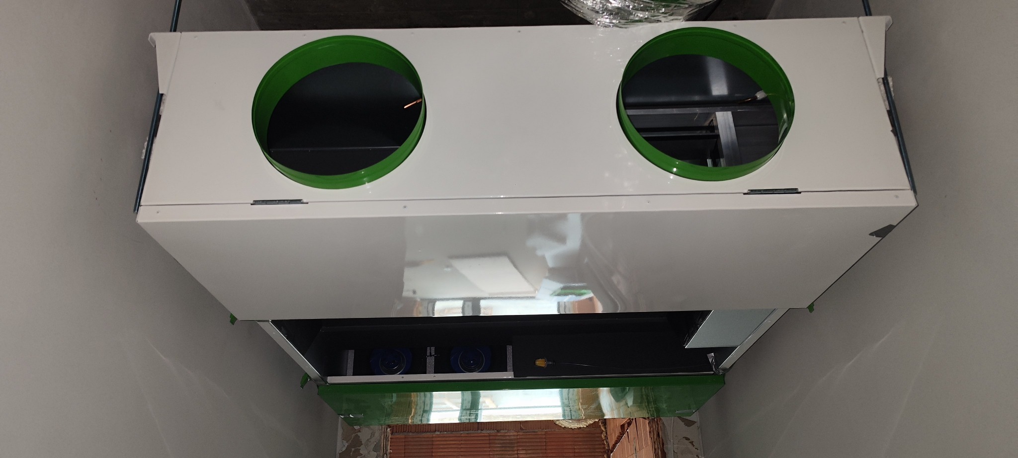 Zamontowany, biały rekuperator z zielonymi otworami wentylacyjnymi, widoczny z dołu, w surowym wnętrzu. Element instalacji wentylacyjnej w trakcie montażu.