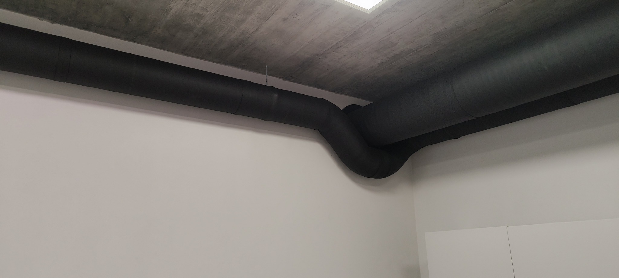Czarne, zaizolowane rury wentylacyjne pod betonowym sufitem w pomieszczeniu. Widoczne łączenia rur.
