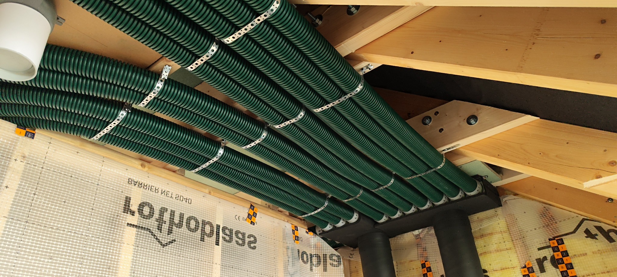 Zielone, karbowane rury wentylacyjne przymocowane do drewnianego stropu za pomocą metalowych obejm. Widoczne elementy konstrukcyjne dachu i folia paroizolacyjna.