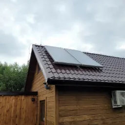 Drewniany domek z brązowym dachem i dwoma panelami słonecznymi na dachu, dodatkowo widoczna klimatyzacja na ścianie budynku.
