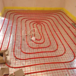 Czerwone rury ogrzewania podłogowego ułożone spiralnie na folii izolacyjnej z siatką, przymocowane plastikowymi uchwytami, widoczne kartony i lampa robocza.