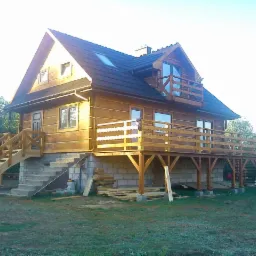 Dwukondygnacyjny dom z bali z drewnianym tarasem na parterze i balkonem na piętrze, widoczne schody wejściowe, fundament z bloczków betonowych, teren wokół domu w trakcie zagospodarowywania.