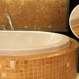 Owalna wanna obudowana mozaiką w odcieniach beżu i brązu, dopasowana kolorystycznie do ściany wyłożonej drobną, kwadratową glazurą. Ujęcie z bliska, z widoczną baterią i słuchawką prysznicową.
