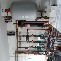 Rozbudowana instalacja hydrauliczna z pompami obiegowymi, zbiornikiem wyrównawczym i licznymi rurami miedzianymi na tle białych płytek.