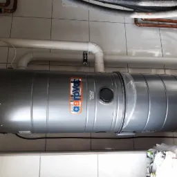 Poziomo zamontowany, szary zasobnik ciepłej wody użytkowej marki Astrovac, z widocznymi rurami instalacyjnymi u góry, na tle białych, kwadratowych płytek ściennych.