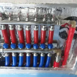 Rozdzielacz ogrzewania podłogowego z czerwonymi i niebieskimi rurami, zamontowany w izolowanej szafce instalacyjnej na tle surowego betonu.