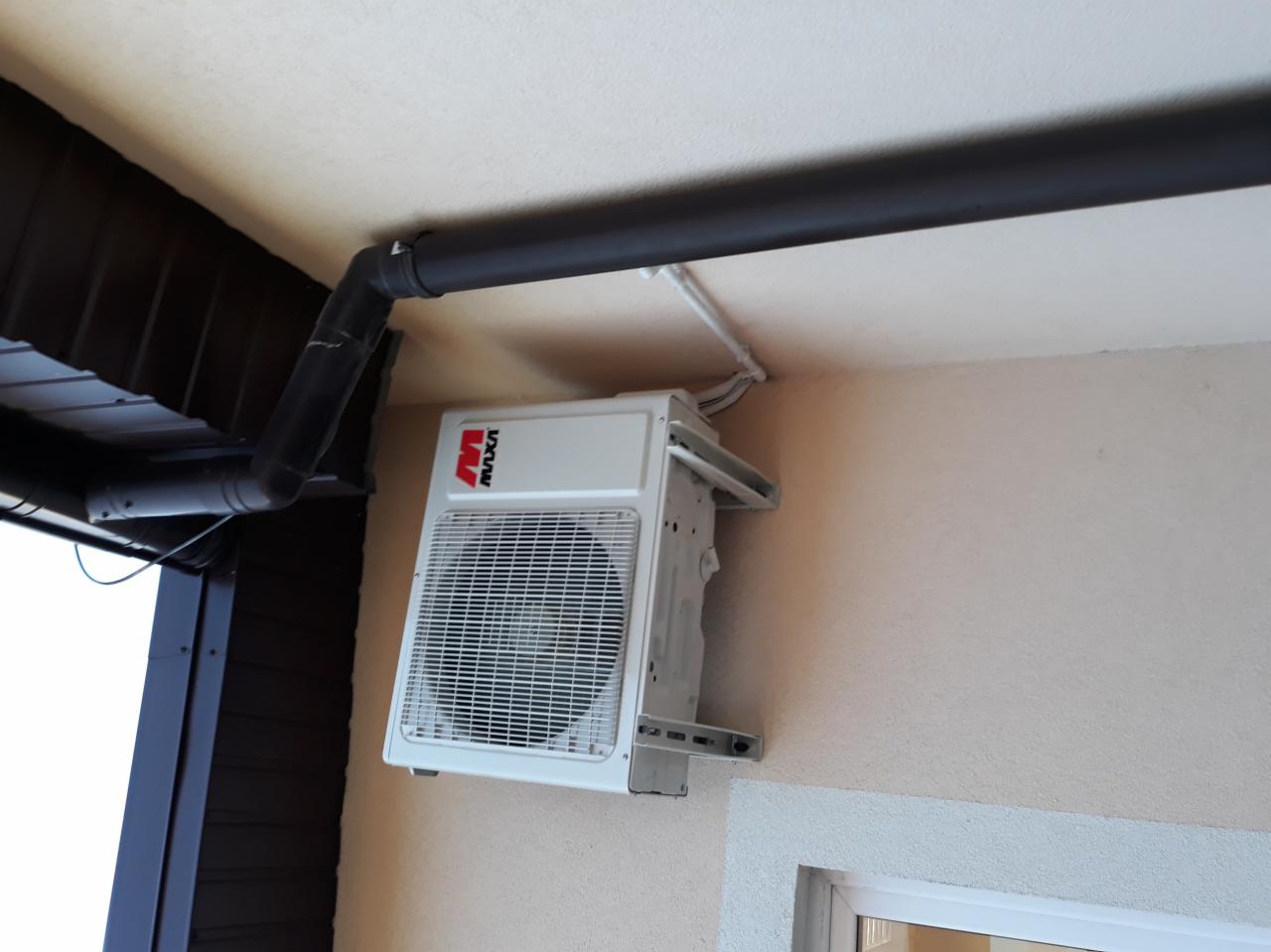Biała jednostka zewnętrzna klimatyzacji marki MAXA zamontowana na beżowej ścianie pod ciemną rynną, widoczne przewody i fragment okna.