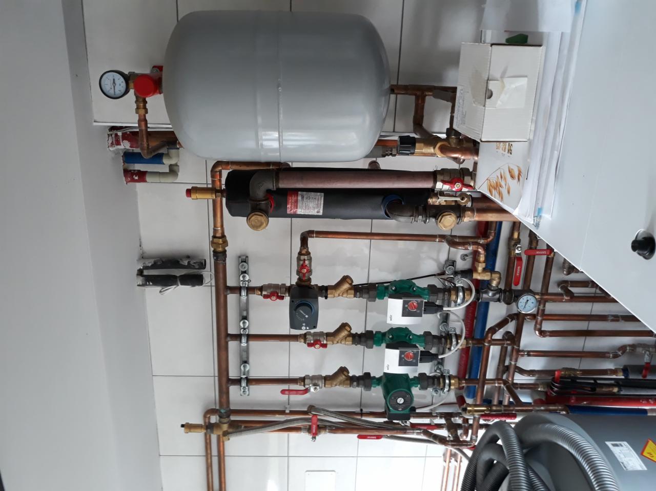 Rozbudowana instalacja hydrauliczna z pompami obiegowymi, zbiornikiem wyrównawczym i licznymi rurami miedzianymi na tle białych płytek.
