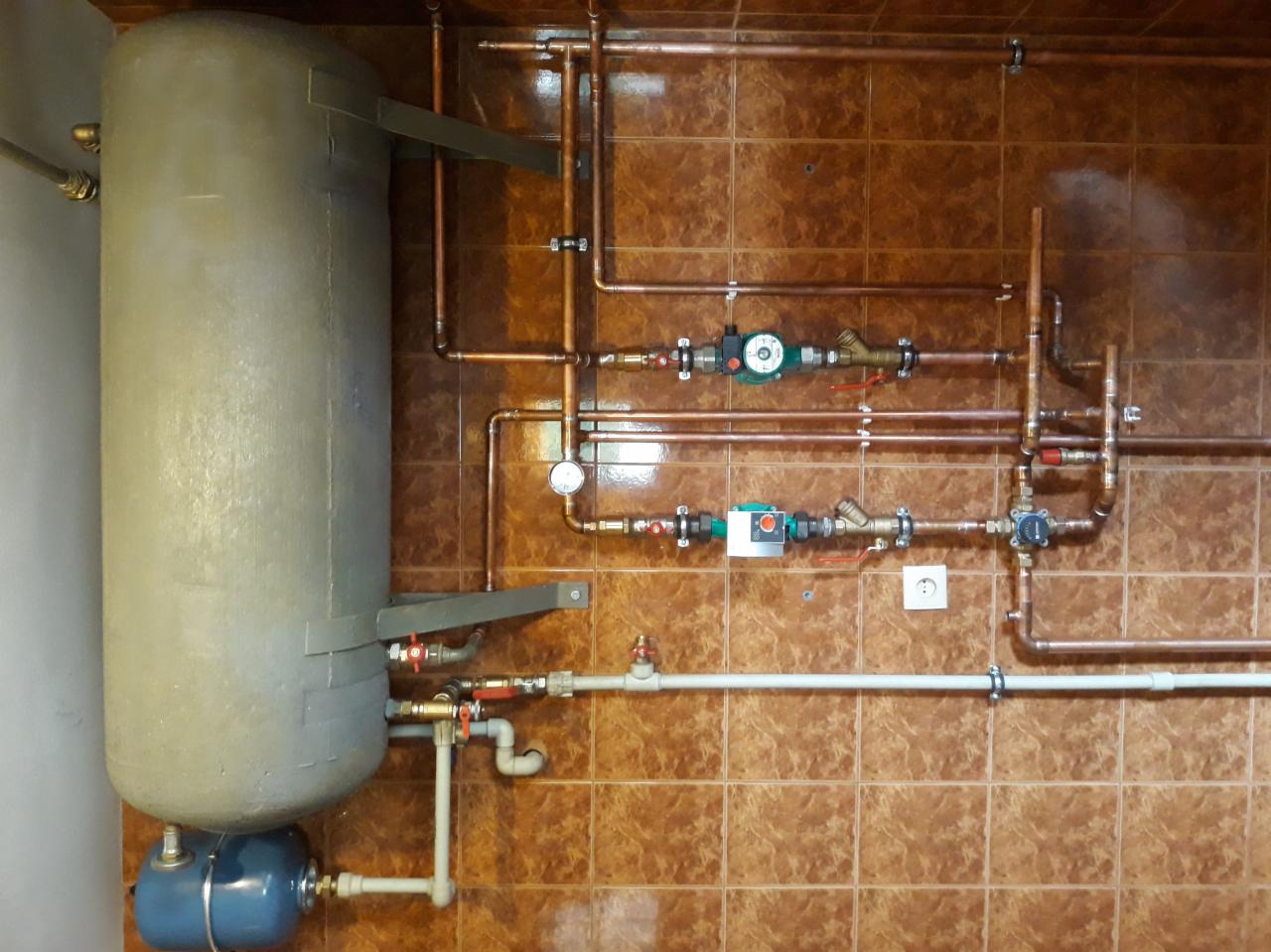 Szary, pionowy zbiornik na wodę z niebieskim zbiornikiem wyrównawczym u dołu, połączony miedzianymi rurami z armaturą i manometrem na brązowej, kwadratowej ścianie z płytek.