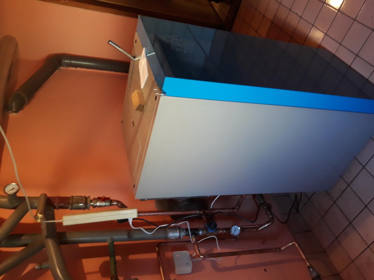 Nowoczesny piec gazowy z niebieskim akcentem, zamontowany na pomarańczowej ścianie z widocznymi rurami miedzianymi i elementami instalacji.