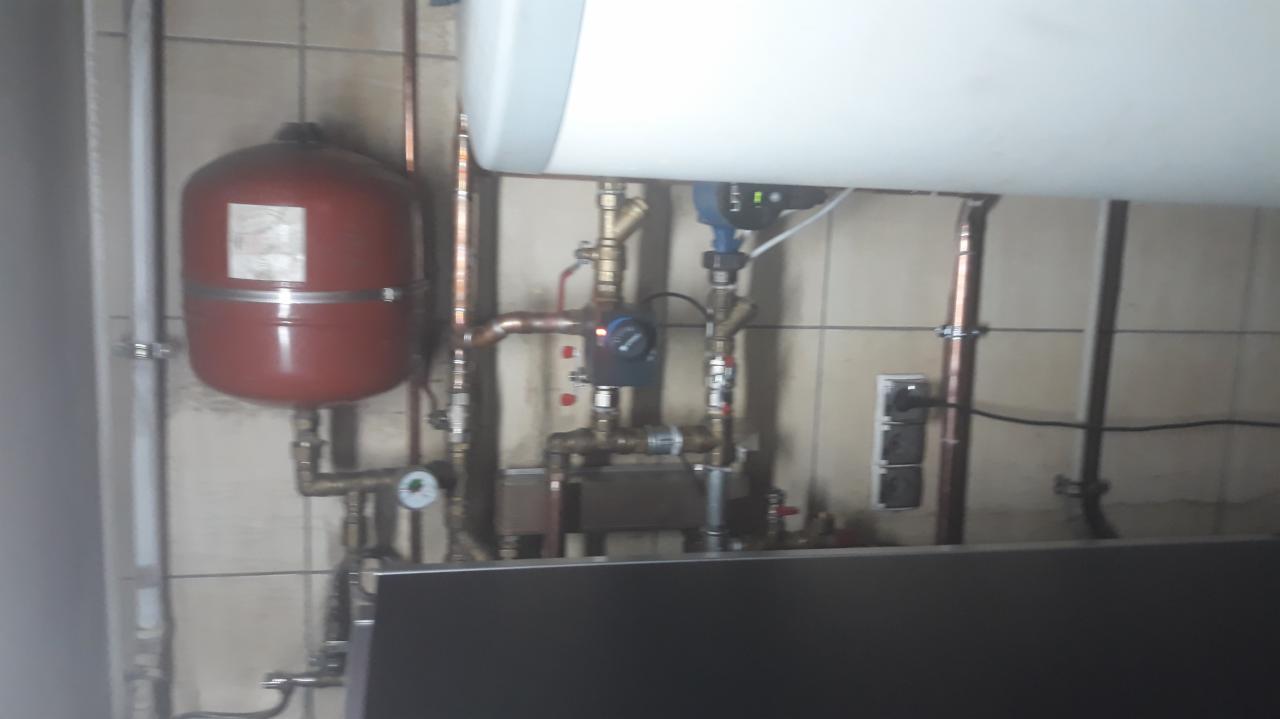 Czerwony zbiornik wyrównawczy ciśnienia, pompa obiegowa i rury miedziane zamontowane na ścianie z płytek ceramicznych, widoczne gniazdka elektryczne.