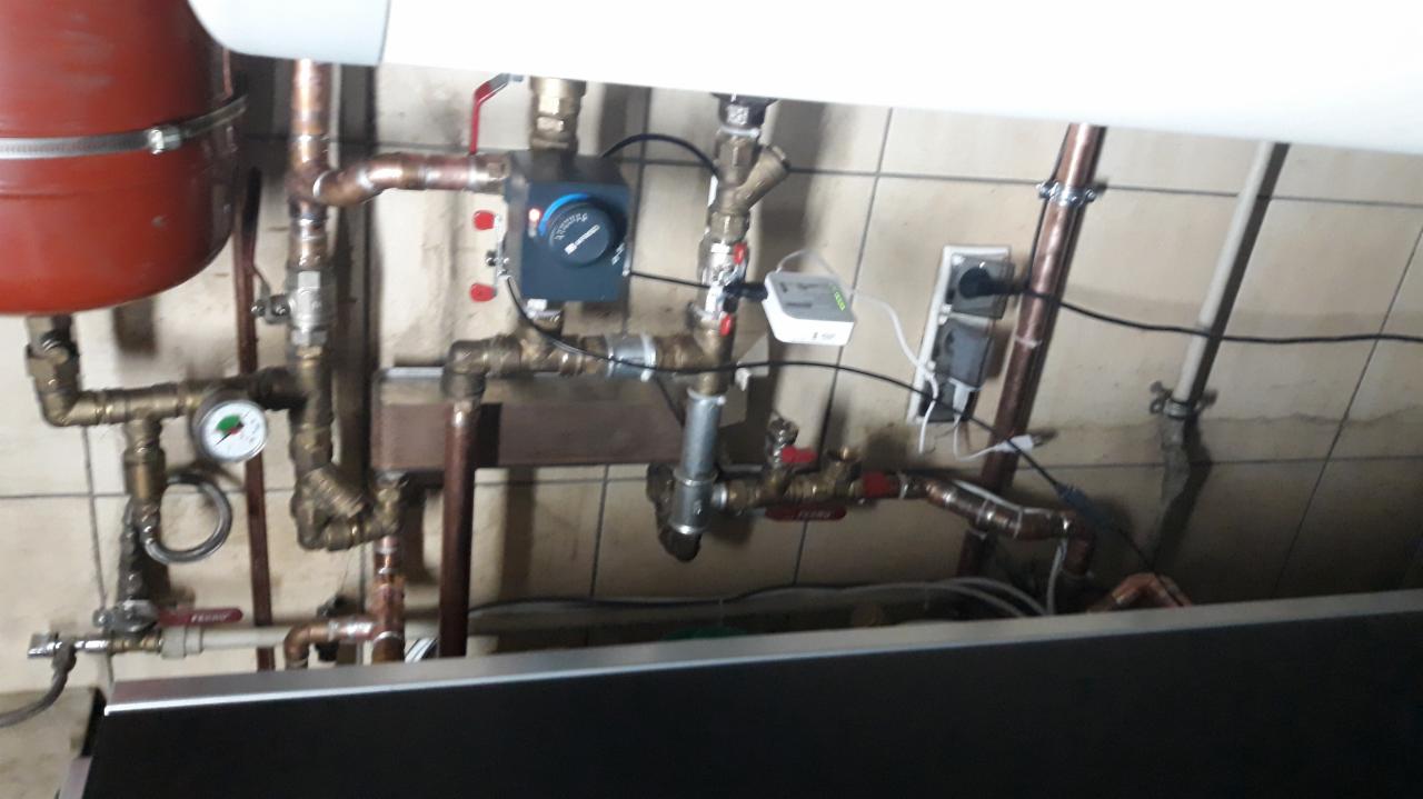 Instalacja kotłowni z widocznym zbiornikiem buforowym, licznymi rurami miedzianymi, manometrem i sterownikiem, podłączona do gniazdka elektrycznego.