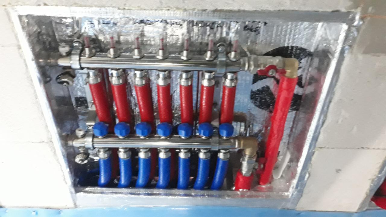 Rozdzielacz ogrzewania podłogowego z czerwonymi i niebieskimi rurami, zamontowany w izolowanej szafce instalacyjnej na tle surowego betonu.