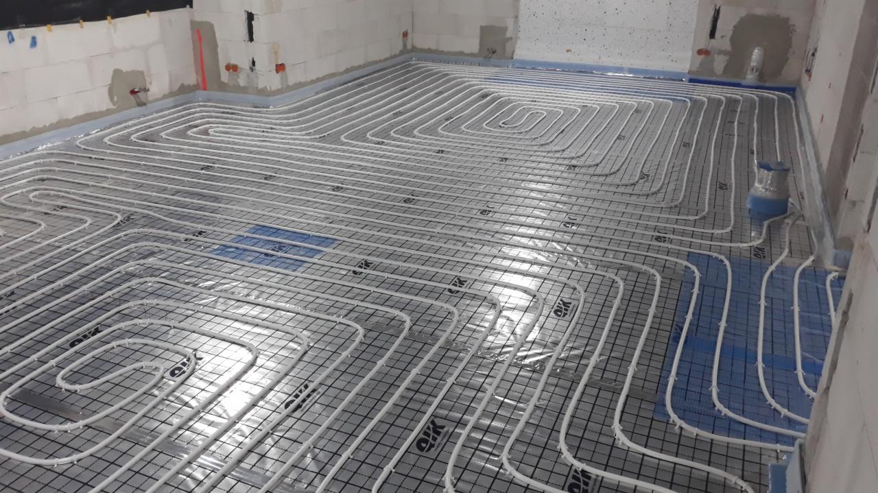 Białe rurki ogrzewania podłogowego ułożone spiralnie na siatce w surowym wnętrzu, widoczne fragmenty ścian z betonu komórkowego i niebieska izolacja przy ścianach.