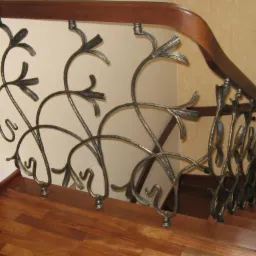 Kuta balustrada schodowa z motywem roślinnym, metalowe elementy w kolorze ciemnego srebra, drewniana poręcz w ciepłym odcieniu brązu, widok z dołu na schody.