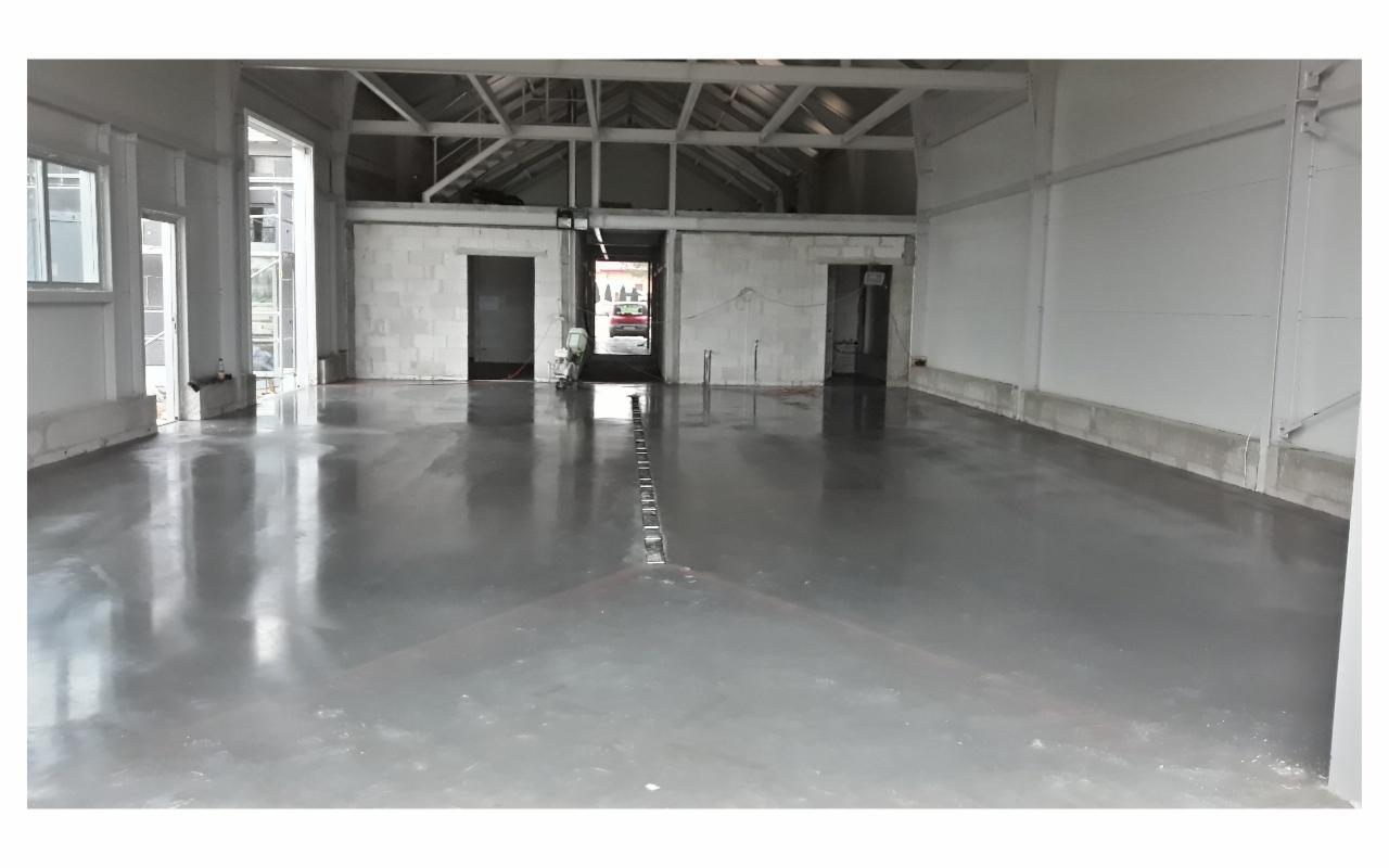 Świeżo wylana, błyszcząca, szara posadzka w surowym wnętrzu hali, widoczne drzwi i elementy konstrukcyjne budynku.