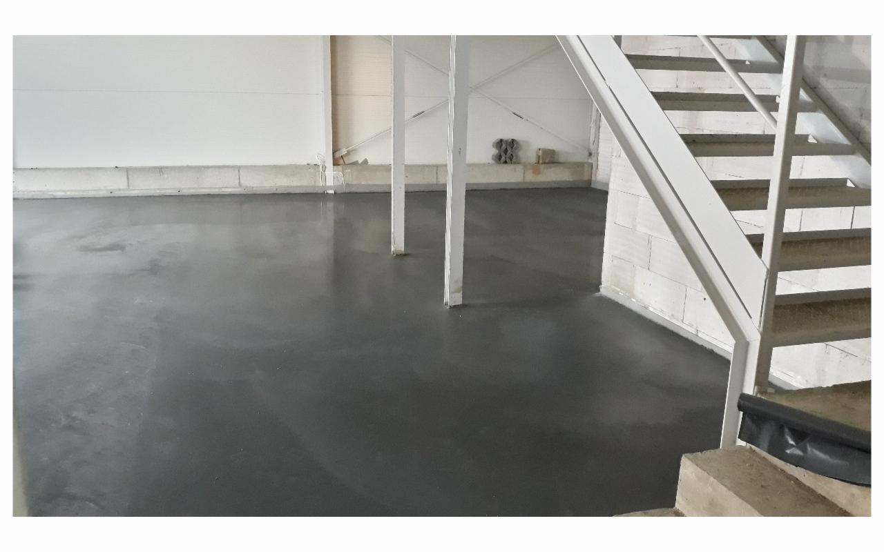 Gładka, szara posadzka w hali przemysłowej, widoczne białe podpory konstrukcyjne i fragment schodów z metalową barierką.