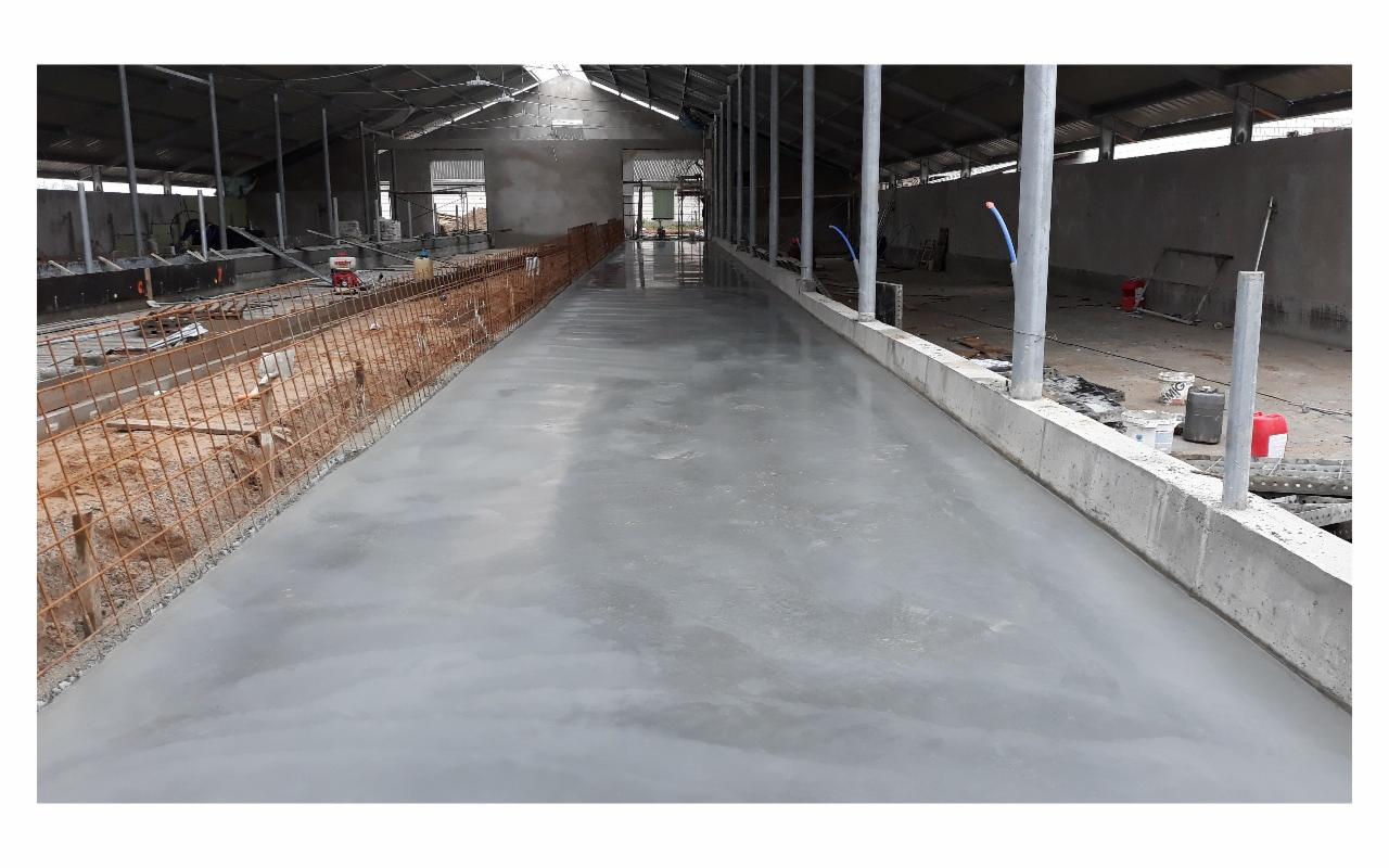 Świeżo wylana, gładka, szara betonowa posadzka w hali przemysłowej z widocznymi elementami konstrukcyjnymi i zbrojeniami po bokach.
