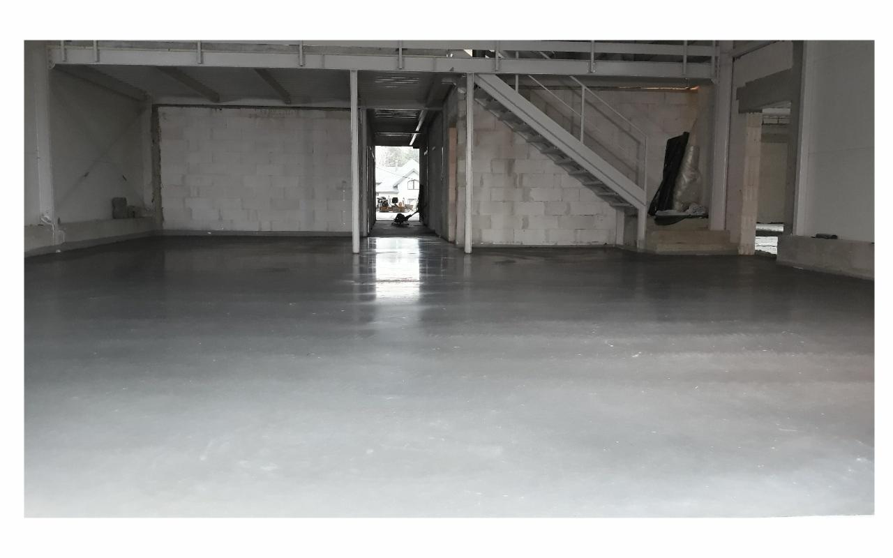 Świeżo wylana, gładka posadzka w surowym wnętrzu hali, widoczne bloczki betonowe ścian i metalowa konstrukcja schodów.