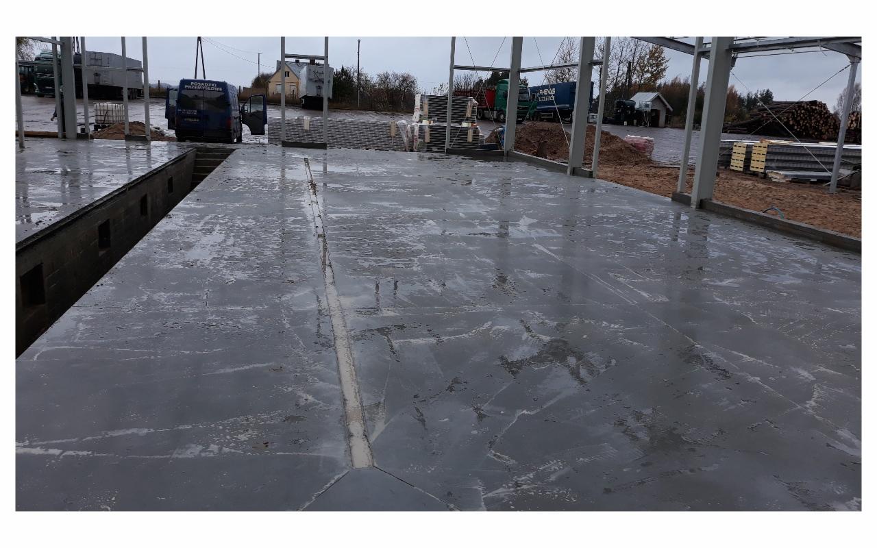 Świeżo wylana, mokra, szara posadzka w hali przemysłowej z widocznymi szczelinami dylatacyjnymi i elementami konstrukcji stalowej w tle.