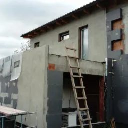 Dom w trakcie prac elewacyjnych: częściowo ocieplony styropianem grafitowym, fragment ściany pokryty szarym tynkiem z widocznym rusztowaniem i drewnianą drabiną opartą o balkon.