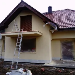 Dom jednorodzinny z nałożonym jasnym tynkiem elewacyjnym, zabezpieczone okna i drzwi folią, drabina oparta o balkon, widoczne przygotowania do dalszych prac.