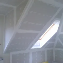 Poddasze z oknem dachowym w trakcie montażu płyt karton-gips, widoczne łączenia płyt i śruby, częściowo zaszpachlowane.