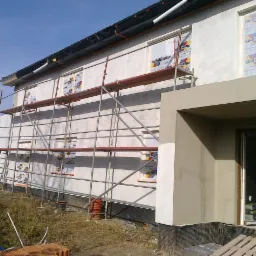Dom w trakcie prac elewacyjnych: rusztowanie, częściowo otynkowana ściana z oknami zabezpieczonymi folią z nadrukiem Austrotherm, widoczne cegły i paleta na ziemi.