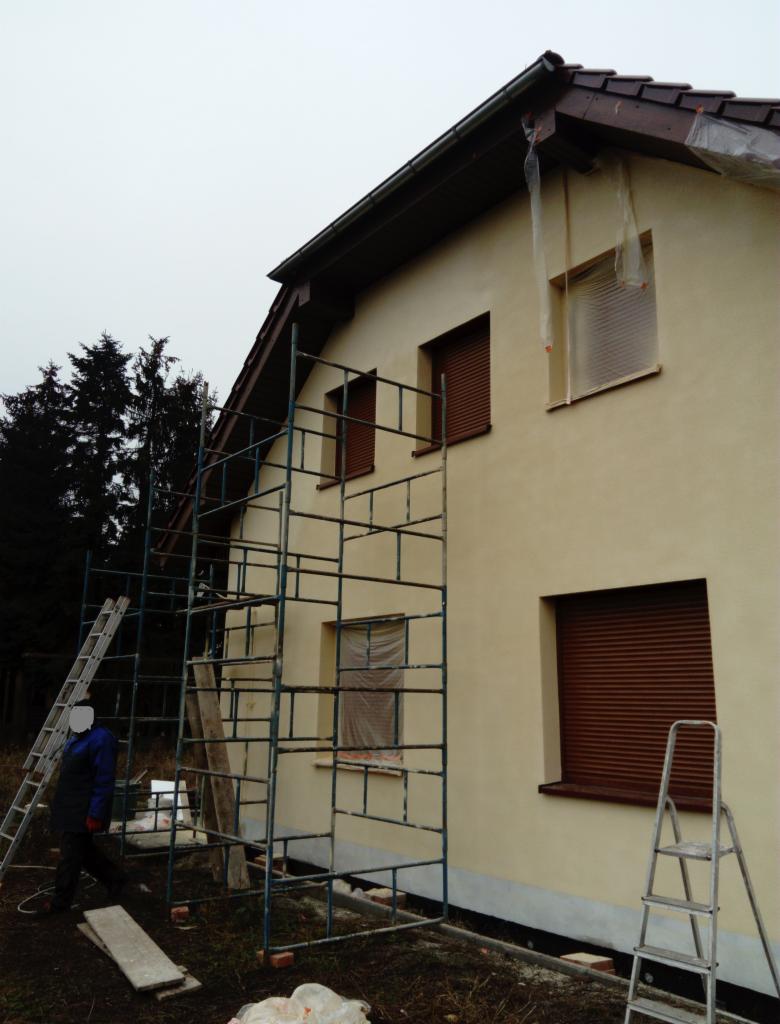 Dom jednorodzinny w trakcie prac elewacyjnych; widoczne rusztowania, okna częściowo zabezpieczone folią, świeżo otynkowana ściana w kolorze kremowym.