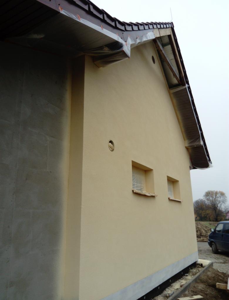 Ściana budynku w trakcie prac elewacyjnych; tynk w kolorze kremowym, okna zabezpieczone folią, widoczny fragment ściany w surowym stanie i fragment dachu z folią ochronną.