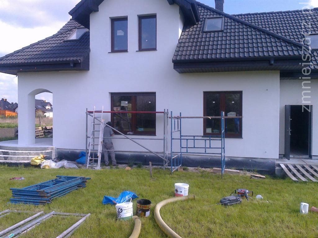 Prace elewacyjne przy domu jednorodzinnym: pracownik na rusztowaniu maluje ramę okienną, widoczne narzędzia, wiaderka z farbą i elementy rusztowania na trawniku.