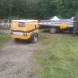 Żółta przyczepa z agregatem na błotnistej drodze, podłączona do przyczepy transportowej z tabliczką informacyjną, ogrodzona taśmą ostrzegawczą.