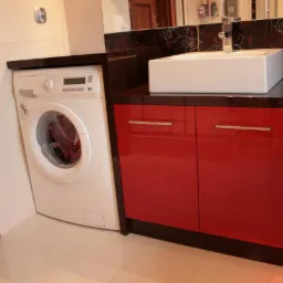 Łazienka z czerwoną szafką pod umywalką, białą umywalką nablatową i pralką obok, płytki ceramiczne na ścianach i podłodze.