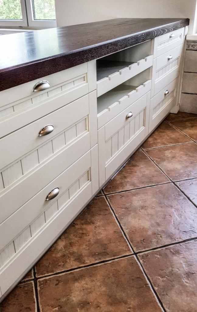 Białe szafki kuchenne z drewnianym blatem i uchwytami w kształcie muszelek, widoczna półka na butelki, brązowa podłoga z płytek.