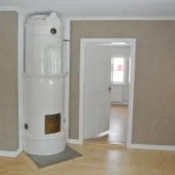 Biały, ceramiczny piec kaflowy w rogu pokoju z jasną podłogą i beżowymi ścianami. Widoczne otwarte, białe drzwi prowadzące do kolejnego pomieszczenia z oknem.